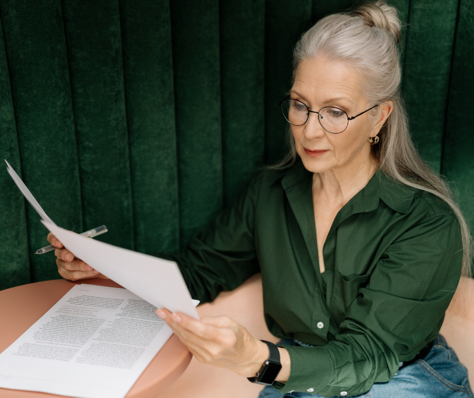 Uma mulher madura olha para diversos documentos em sua mesa de trabalho enquanto segura uma caneta esferográfica preta. A senhora tem cabelos brancos, usa um óculos preto redondo, uma elegante camisa verde e uma calça jeans.