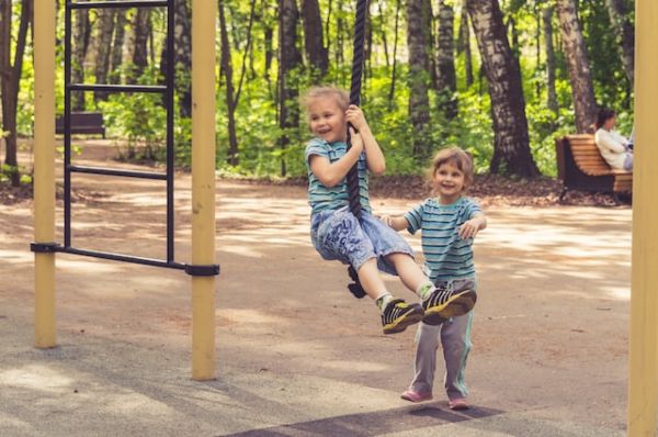 one child swings on a rope swing while another child stands behind them watching and smiling