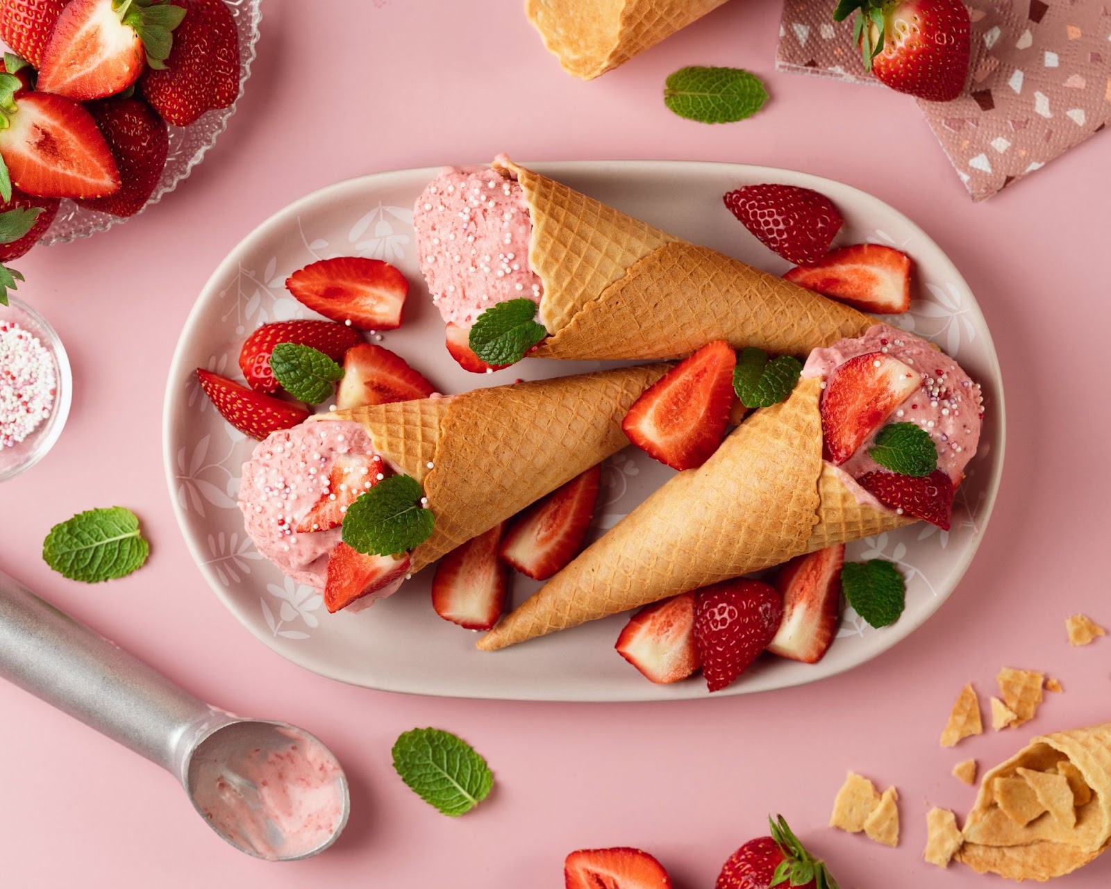 a table display of three ice cream waffle cones with strawberry ice cream, surrounded by strawberry halves and mint leaves on a pink surface