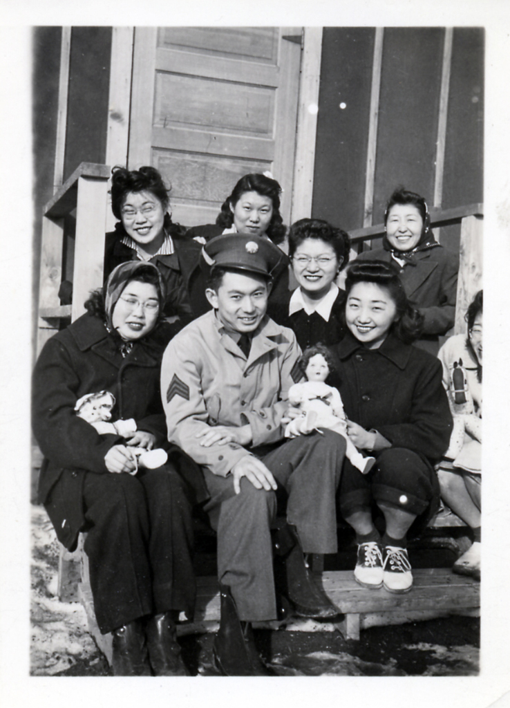 Nisei soldier Tak Kondo sitting on the front steps of a barrack in Heart Mountain with a group of women.