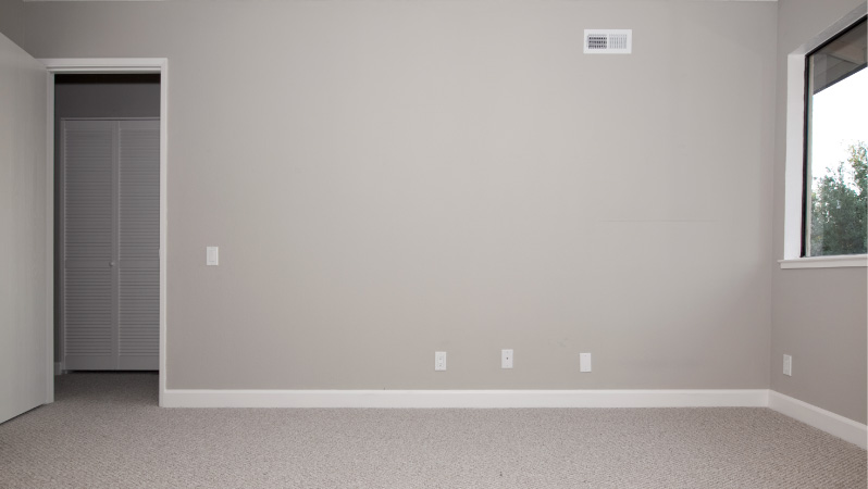 An unused bedroom in a home — which wastes energy, money, and other resources — with blank gray walls and gray carpet.