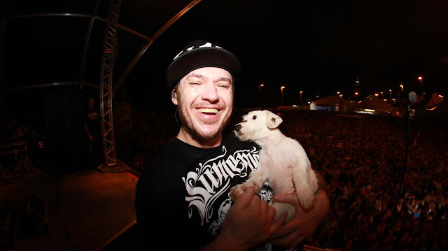 Chorão tem nos braços um cachorro. Ambos estão em cima de um palco onde a banda Charlie Brown Jr se apresentou. Atrás dele há uma vastidão de pessoas.