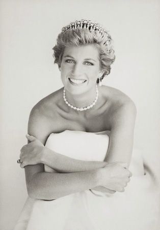 Princesa Diana está sentada aparentemente no chão, usando um vestido branco longo, seus braços se entrelaçam. Em sua mão direita usa um anel. Seu cabelo é curto e está penteado para trás. Ela sorri. Usa adereços no cabelo, uma tiara de princesa e um colar de pérolas.