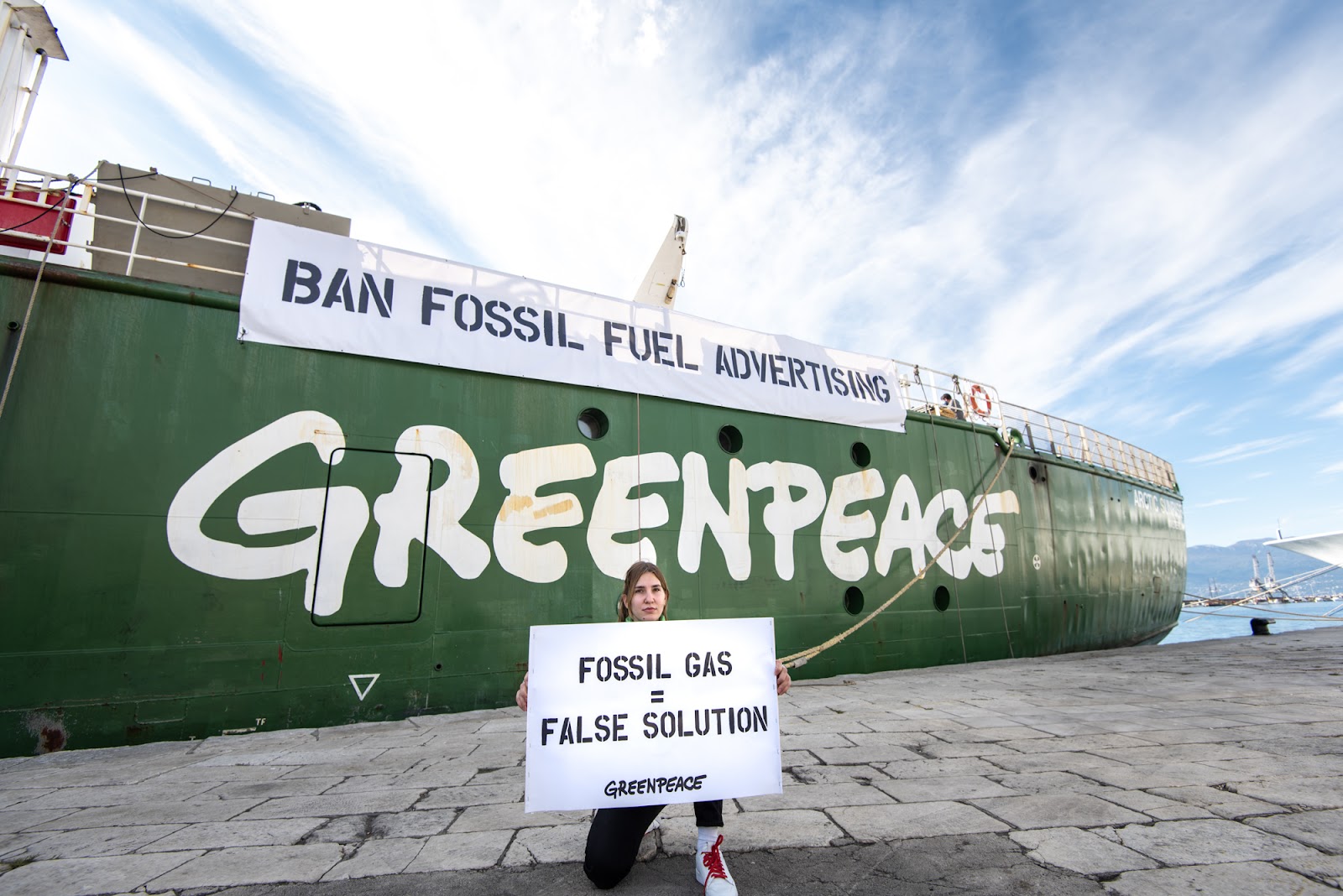 Greenpeace ship Arctic Sunrise displays a banner from the deck reading Ban Fossil Fuel Advertising. Activist on the dock in front of the ship holds banner saying Fossil Gas = False Solution. 