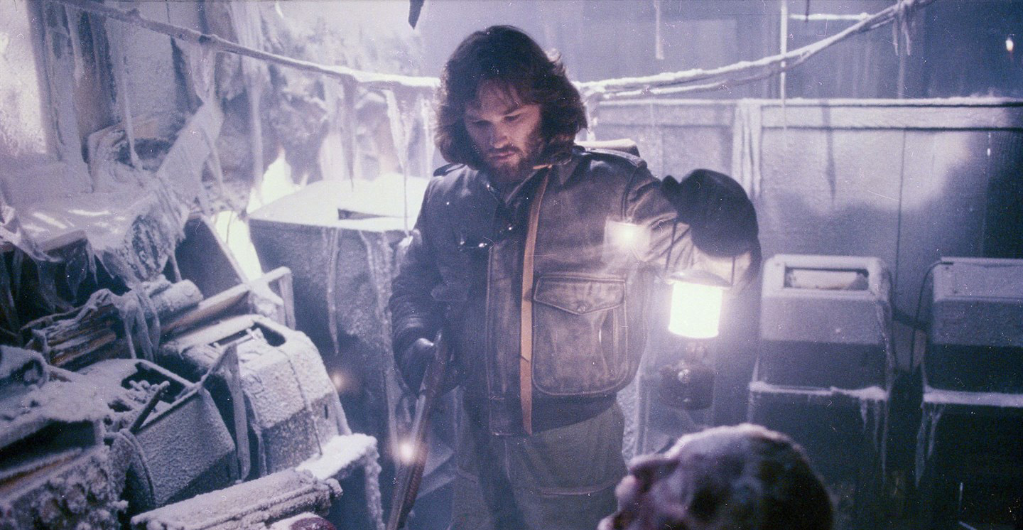 A still of MacReady, played by Kurt Russell, looking down at a frozen corpse. He is holding a gun and a lantern.