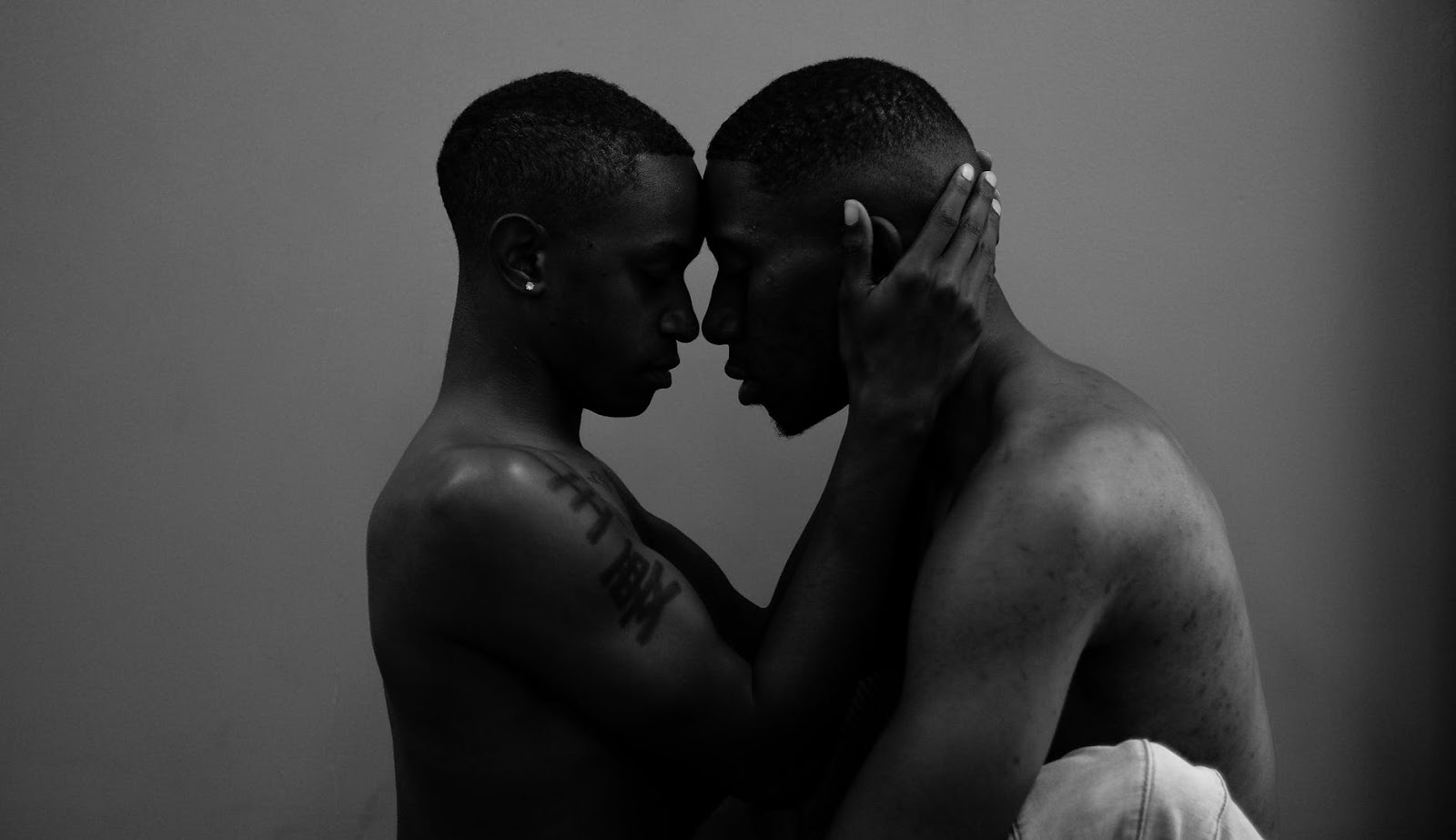 Two shirtless men embracing each other. Their faces are facing each other.