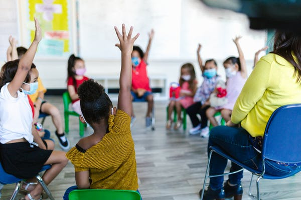 Preschoolers raising their hands sitting in a circle with their teachers. 
