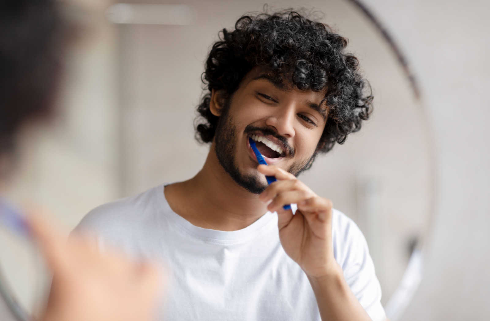 A man with curly hair in a white shirt brushing his teeth in front of a mirror.