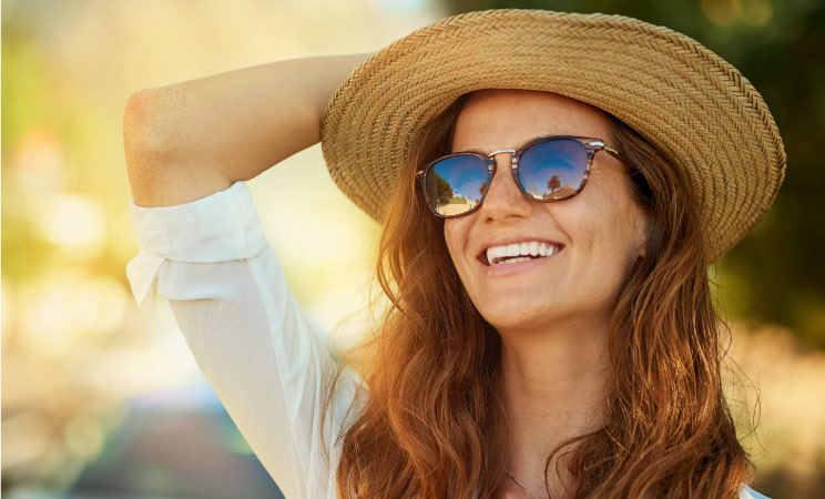 A smiling woman is wearing sunglasses and a straw hat and looking up on a bright sunny day. 