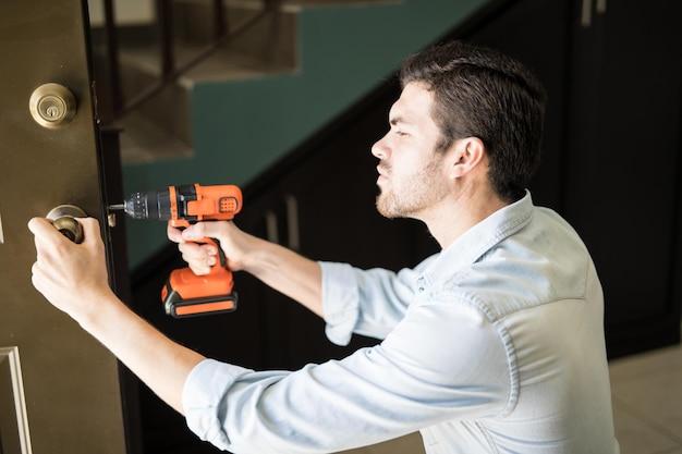 Vista de perfil de um trabalhador braçal atraente usando uma furadeira elétrica para consertar uma maçaneta de porta em uma casa coberta na Assistência Flex Domiciliar