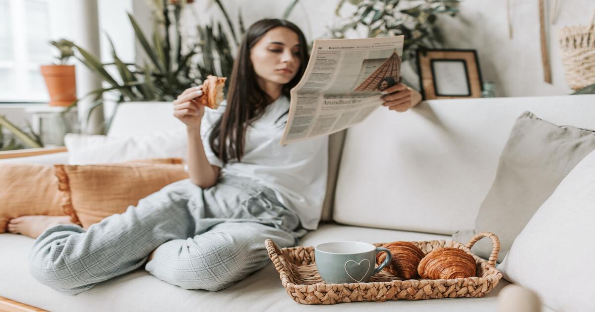 Small business holiday gift guide–An image of a woman in loungewear sitting on a white couch, reading a newspaper, and eating a croissant with a tray of croissants and a cup of coffee beside her. 