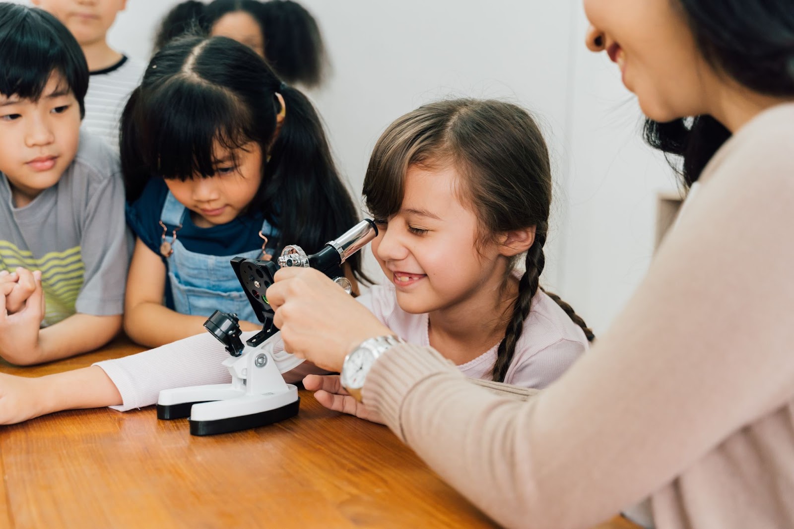 Menina olhando por um microscópio com auxílio da professora e colegas em sua volta, exemplificando uma equipe interdisciplinar