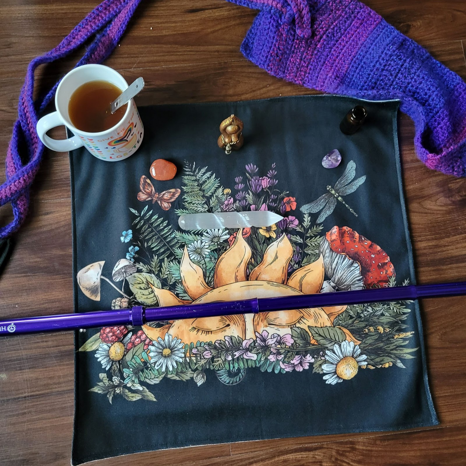 Image description: a purple walking cane is laid across a tarot cloth decorated with flowers, mushrooms, and a rising sun. Above the cane is laid out a selenite wand, carnelian and amethyst stones, a Hestia statue, a mug of tea, a smaller apothecary bottle, and a purple crocheted cane holder.