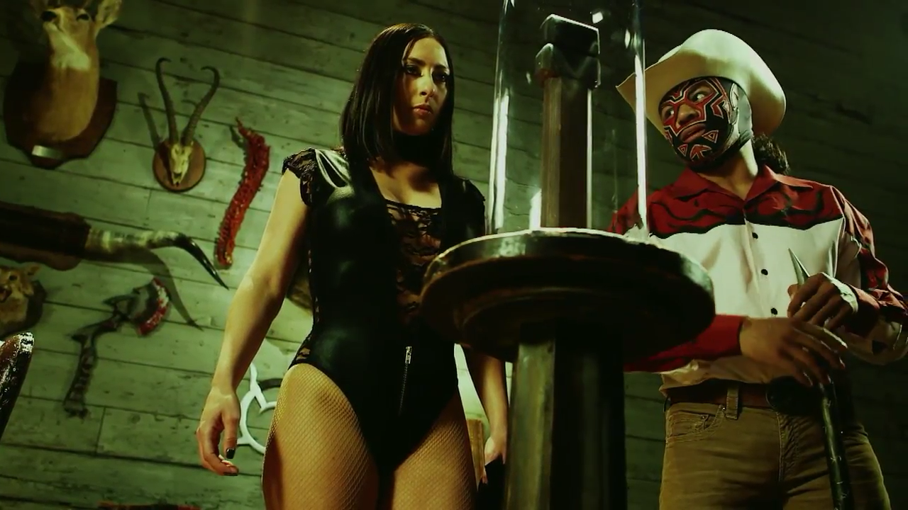 Screen cap from Lucha Underground. Catrina, a dark haired woman wearing a black leotard, is glaring at an empty glass case that used to hold a steel gauntlet. King Cuerno, a main in a red and black mask wearing a wide brimmed hat and black and red clothes, looks knowingly at her. They are surrounded by Cuerno's tropies, mostly the heads of animals mounted on the wall behind them. 