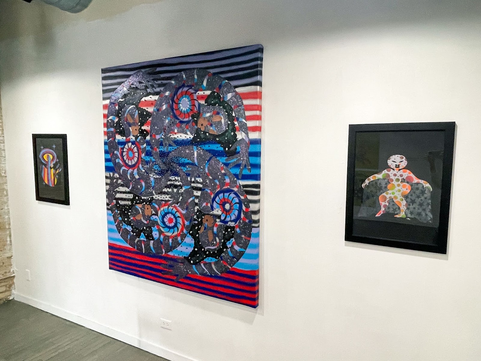 Image: An installation view of won't you celebrate with me with three pieces in a row. The middle piece is "Spinning Satellites" by Erin LeAnn Mitchell. It is a large, colorful abstract piece with horizontal stripes in the background and swirls in the foreground. The two pieces on either side of it are of colorful figures on a black background. Courtesy of the artist and FLXST Contemporary.