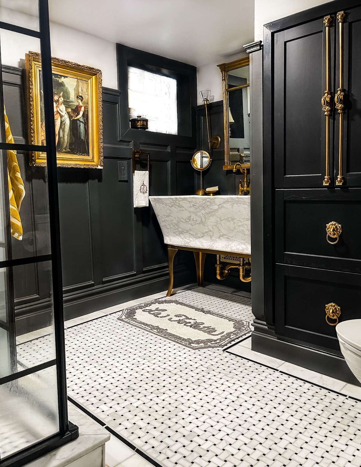 Ashley's traditional black and white colored bathroom is styled with an oil painting, marble sink, and a large French gilt mirror