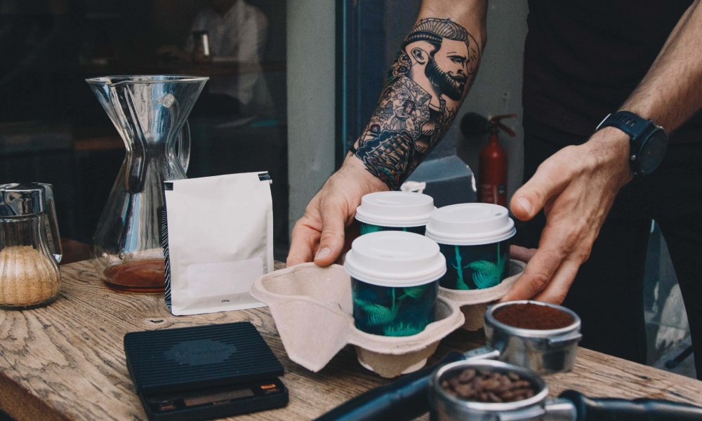 Caucasian male with tattoos places three takeaway coffee cups on wooden table surrounded by coffee brewing equipment and packaging. 