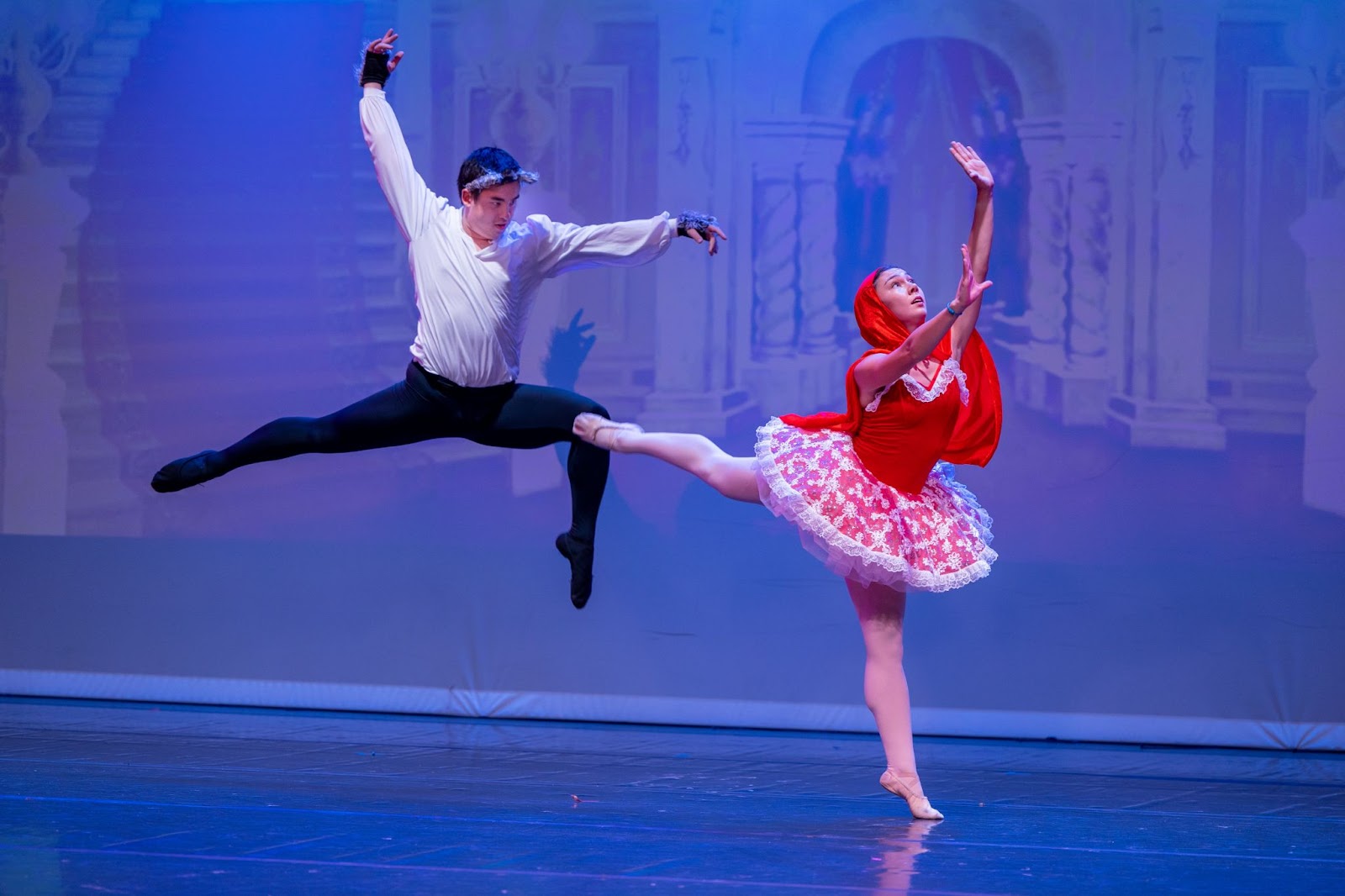 On stage, a male dancer hangs midair with his legs flexed. A female dancer poses beside him in a red shawl and tutu, holding her hands toward the sky.