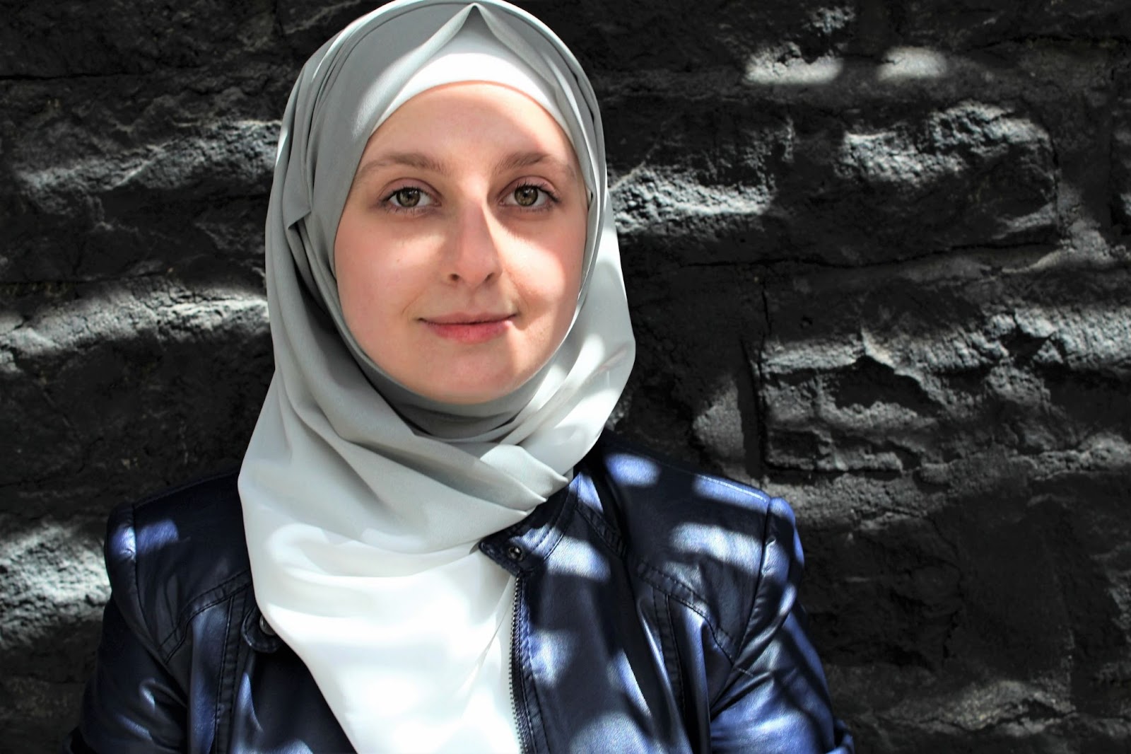 head and shoulders shot of Salwa waring a light grey and white scarf and black jacket , stood against a dark grey brick wall. 