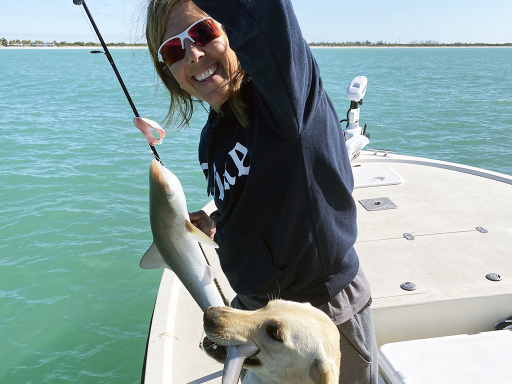 A dog bites a fish over the side of a boat after a woman has caught it fishing