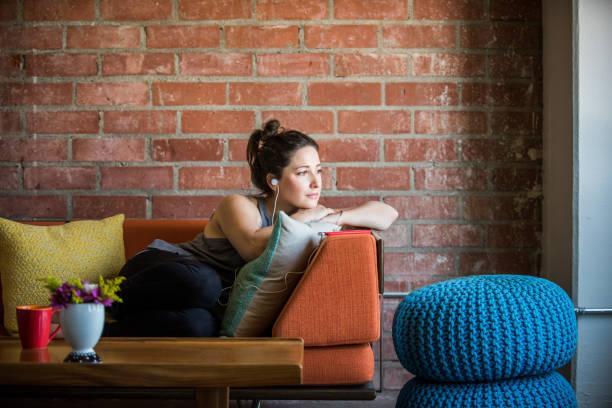 Young Woman Intently Listening to a Podcast Young caucasian woman intently listening to podcast, being reflective and introspective while lying on her side on her sofa in her loft apartment audio book stock pictures, royalty-free photos & images