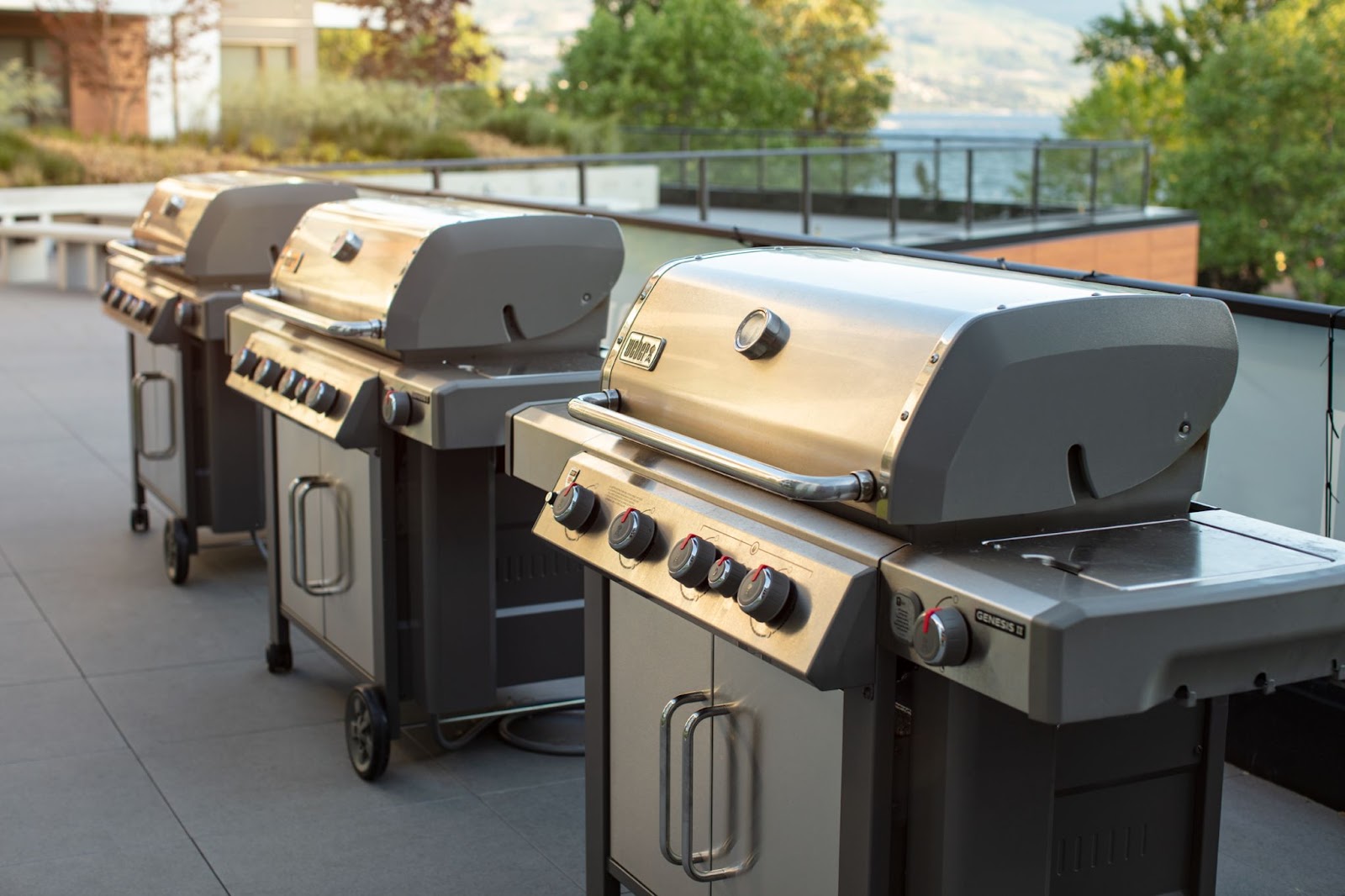 three Weber grills on the third floor terrace of The Shore Kelowna, with the lake and mountains and trees in the background