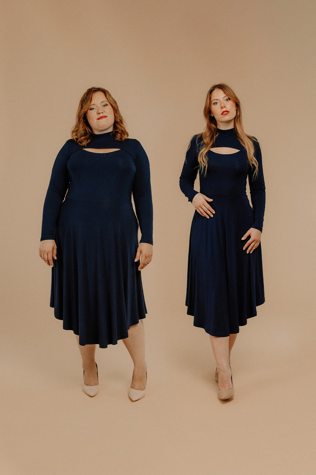 A photo of two models side by side.  The left model is plus sized, the right model is a medium build.  Both are fair skinned with red hair that is long.  The dress they are wearing is navy with a skirt that is longer at the center front than at the sides.  It has sliver of skin exposed at the high chest.  The long sleeves are close fitting.