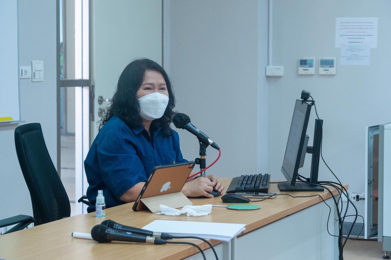 A person wearing a mask and sitting at a desk with a microphone Description automatically generated with medium confidence