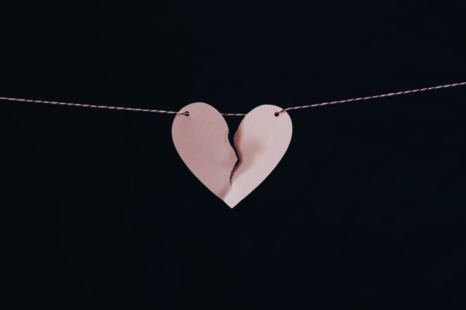A pink, paper heart with a tear halfway down the middle, threaded onto a red and white string. This is all against a plain black background.