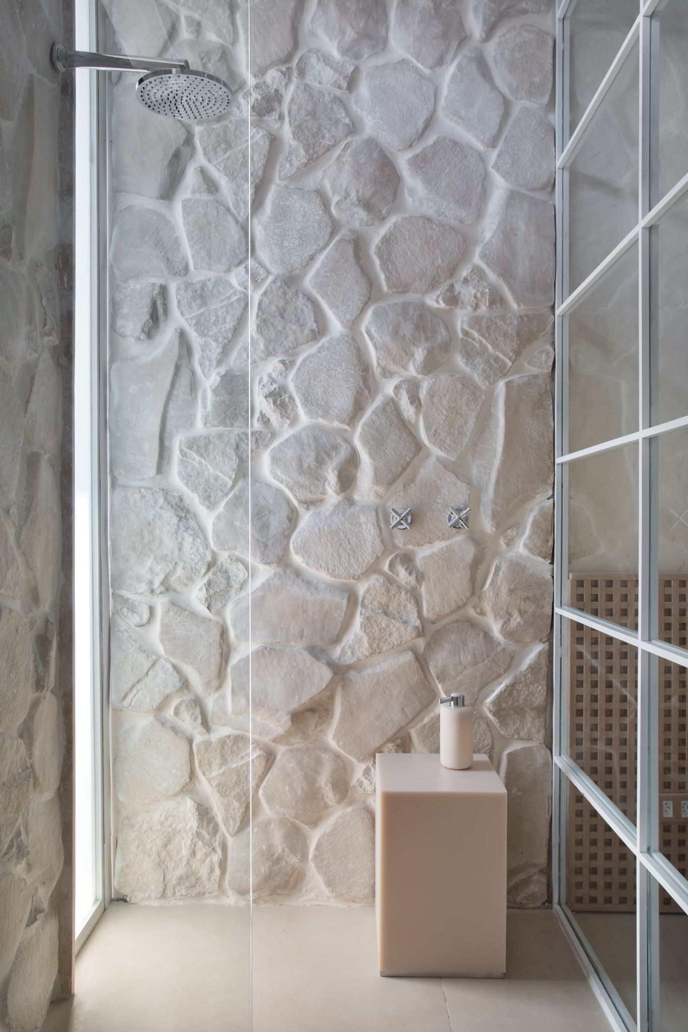 Box de banheiro com parede revestida de pedras brancas, porta de vidro do box com molduras branca e piso branco