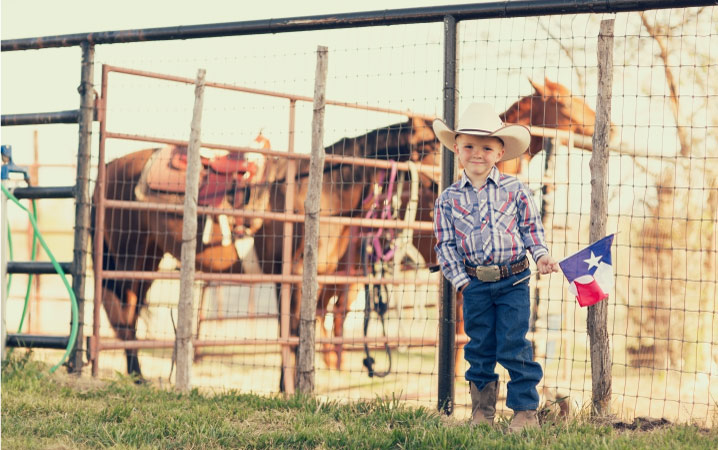 A little boy dressed in Western attire is smiling and holding a small Texas state flag beside a horse paddock. Two horses equipped with Western saddles are standing behind him. 
