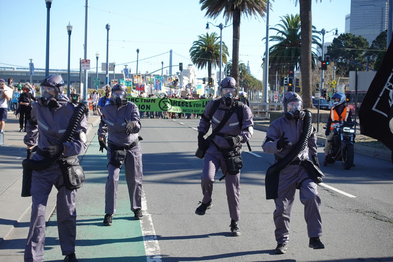 Rebels in miltary dress and gas masks march rigidly as part of a large rally in sunny San Franciso bay.