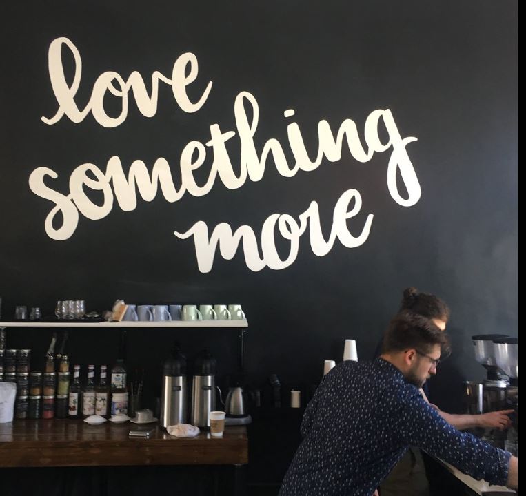 Image of baristas making coffee with a black wall behind them that says lvoe something more