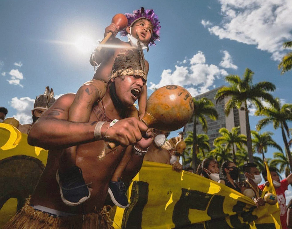 An indigenous man cheers as his small boy sits on his shoulders. Both hold maraccas.