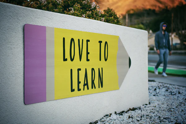 A white short wall with a large image stuck on it of a big yellow pencil with a purple eraser and the words LOVE TO LEARN written on it.