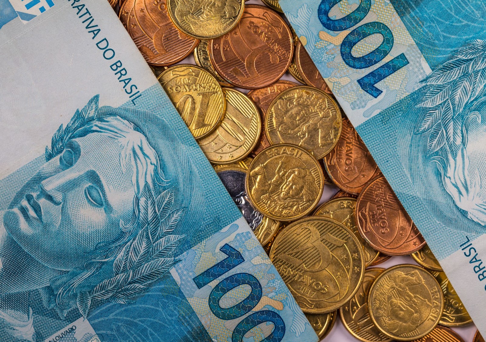 Imagem de duas cédulas de R$ 100,00 e diversas moedas de 5, 10 e 25 centavos.