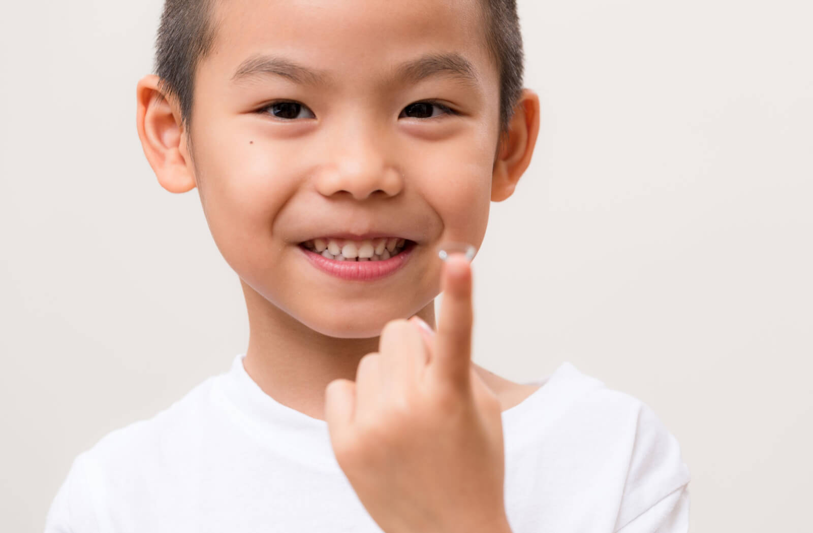 A male kid in a white is smiling and holding a contact lens on the tip of his finger.
