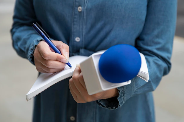 mão feminina branca segurando um microfone e um caderno enquanto faz anotações no que parece ser uma reportagem em curso