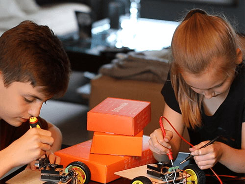 Two children concentrate as they work to assemble the WIFI Robot from one of Thimble's STEM kits at a table. The table is covered in orange Thimble delivery boxes.