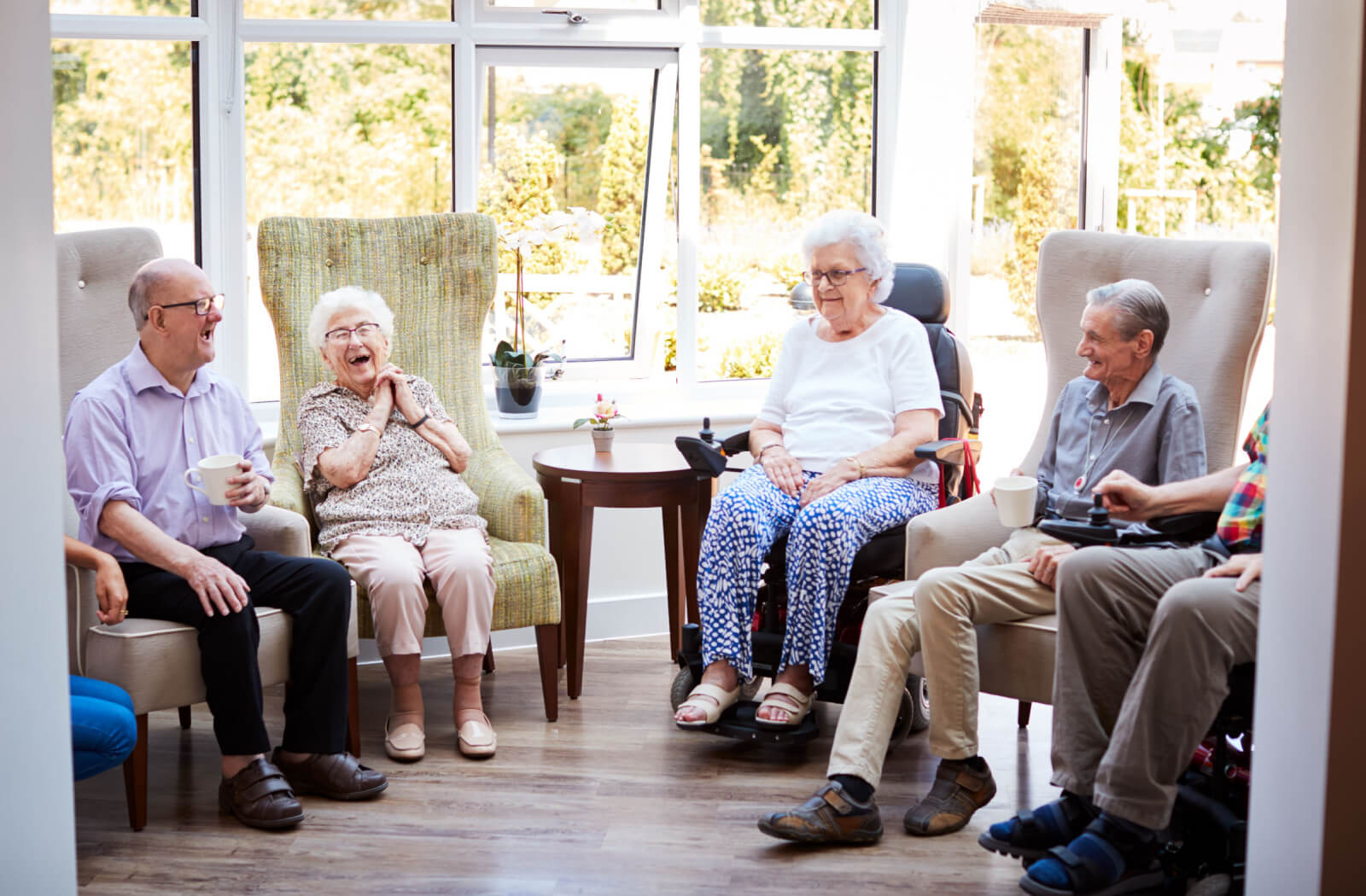 A group of seniors sitting in a semicircle in a common area, drinking tea or coffee and laughing.