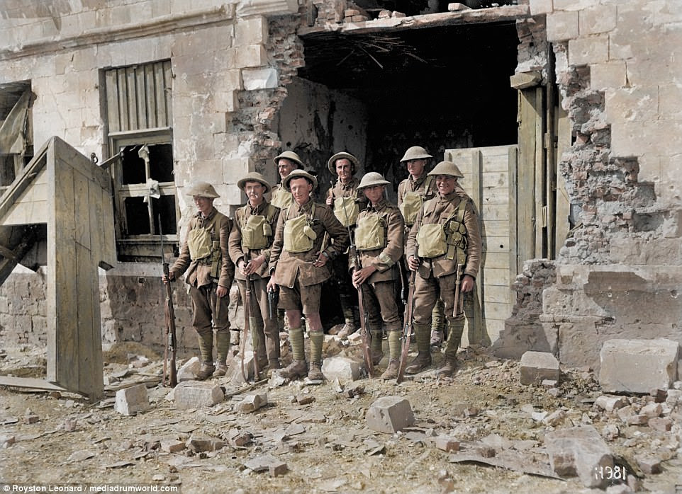 A group of eight British soldiers stand next to a blown-out building. They smile as they pose for the camera wearing their helmets