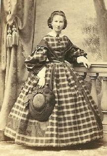 1861-1865, Lovely plaid dress with really full sleeves & undersleeves; solid sleeve caps that extend to bodice center like low lapels and a wonderful hat! Love this design. (The small dainty figure helps!)