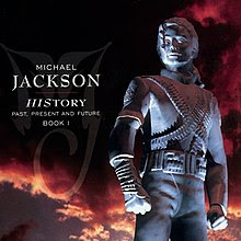 A image of a silver status that is wearing a military-like outfit and has its hair clipped behind its head. To the left of the statue the words "MICHAEL JACKSON" are written in white letters and underneath those two words are other words written in smaller white print. Behind the statue, a sky with clouds that are black and red can be seen.