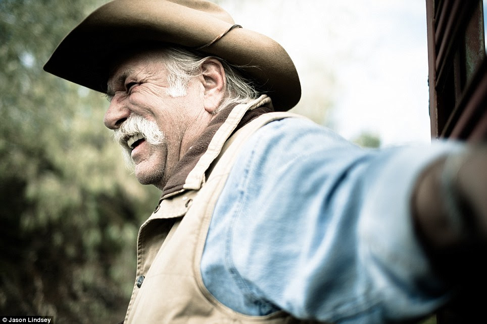 A rancher with a snow-white mustache smiles while leaning against a wall
