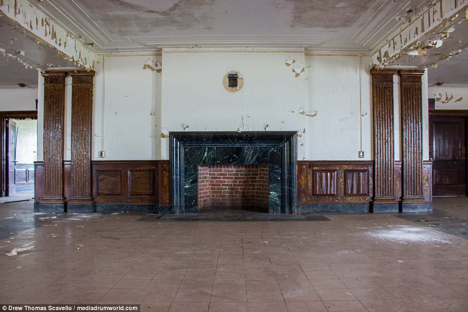 A fireplace has been cleaned and emptied to reveal the red brick finish behind is while the wooden paneling appears badly damaged and pieces of the ceiling have crashed down to the floor