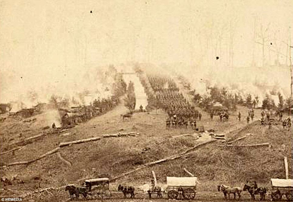 Fog of war: Three horse-drawn covered wagons trundle past soldiers marching in formation between rows of small cabins and tents.