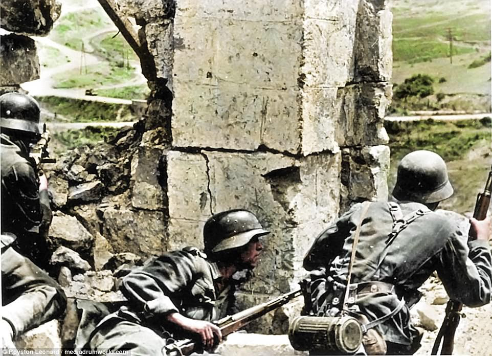 Nazi soldiers in action towards the end of World War Two. Hiding behind a battered wall or archway, they take cover from British and allied forces. One soldier, left, points his gun at what appears to be an army vehicle making its way up the dusty road