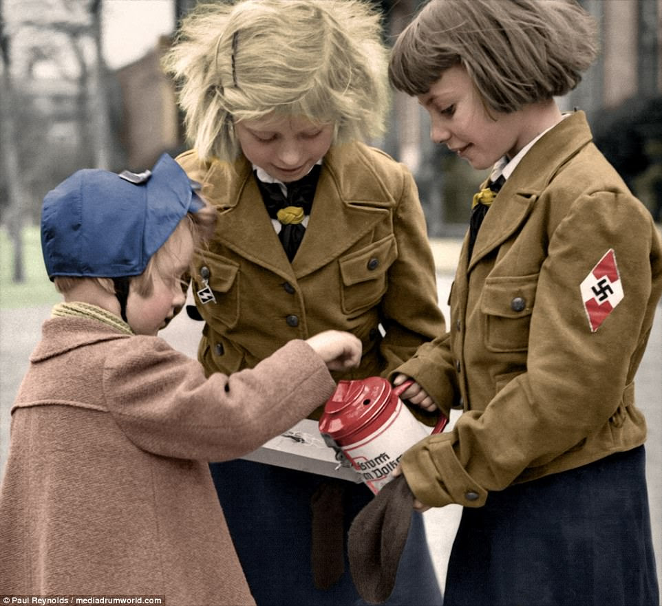 Two girls from The League of German Girls - known in Germany as the Bund Deutscher Mädel (BDM), part of the Hitler Youth - wear smart jackets with Nazi badges as they collect donations from another child for the German Winter Relief Organisation