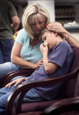 Photograph of a mother sitting in a doctors waiting room with her son who has hurt his nose
