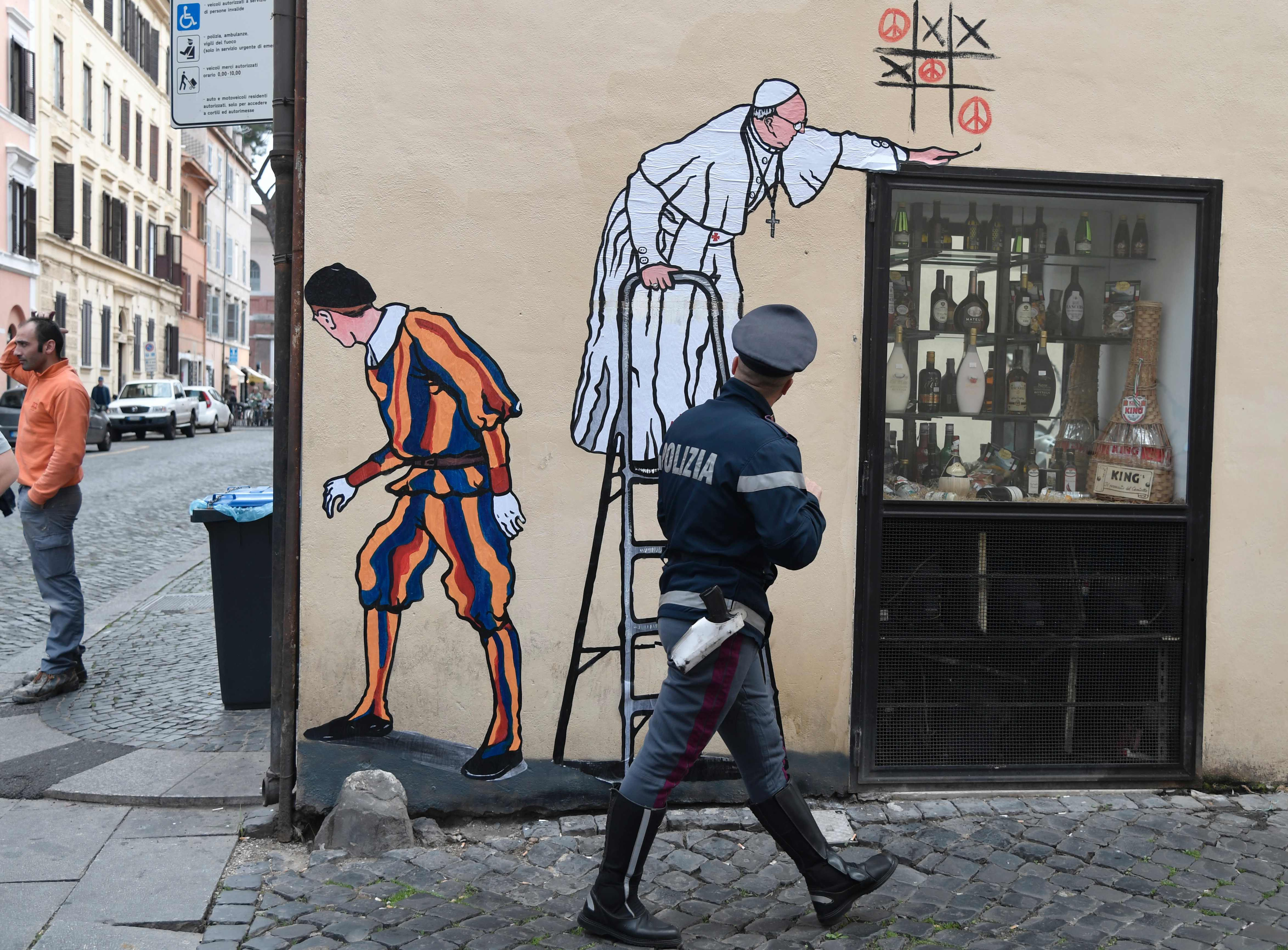 A policeman watches a new street-art collage by Italian artist Maupal showing Pope Francis playing tic-tac-toe and drawing peace signs as a Swiss guard keep watches the street near the Vatican on October 19, 2016 in Rome. / AFP PHOTO / TIZIANA FABITIZIANA FABI/AFP/Getty Images